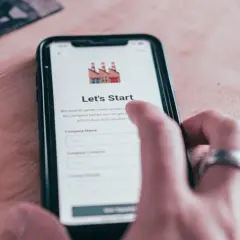 Dłoń z pierścionkiem dotyka ekranu smartfona z aplikacją "Let's Start". Aplikacja zbiera dane firmy, wysyłając powiadomienia.