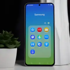 Telefon Samsung z ikonami aplikacji, w tym Bixby. Obok stoi zielona roślina w białej doniczce.