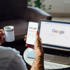 Mężczyzna z kubkiem kawy przegląda telefon, na którym widać stronę Google. Obok stoi laptop z otwartą wyszukiwarką. Chce wiedzieć, jak wylogować się z konta Google.