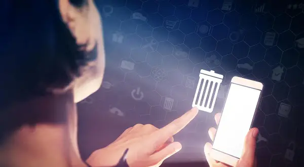 Dłoń dotyka ikony kosza na śmieci na ekranie smartfona, symbolizując czyszczenie telefonu i porządkowanie danych.