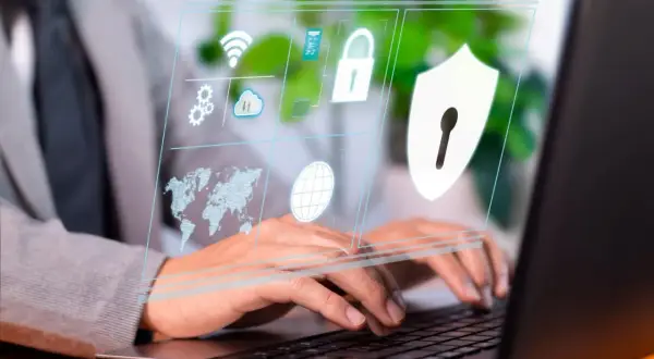 Osoba pracuje przy laptopie, a na ekranie wyświetla się interfejs z ikonami symbolizującymi cyberbezpieczeństwo: globus, mapa świata, chmura, kłódka i tarcza.