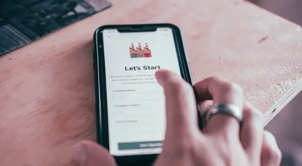 Dłoń z pierścionkiem dotyka ekranu smartfona z aplikacją "Let's Start". Aplikacja zbiera dane firmy, wysyłając powiadomienia.