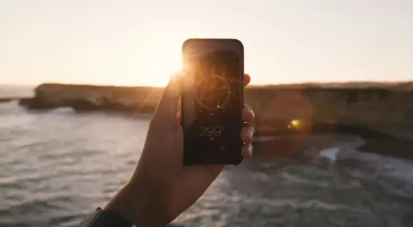 Dłoń trzyma telefon z włączonym kompasem w tle zachodzącego słońca nad morzem.