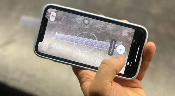 Dłoń trzyma smartfon, na ekranie którego działa aplikacja miarka. Wirtualna linijka mierzy odległość na powierzchni.