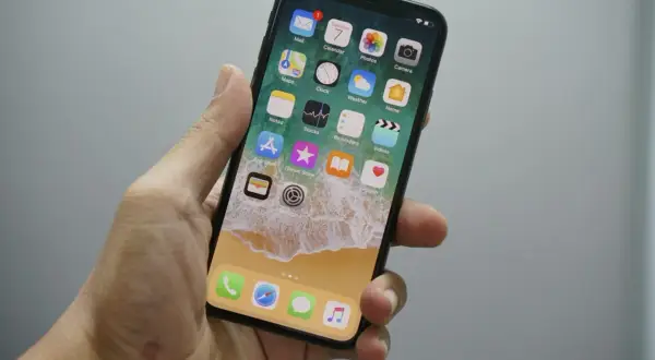 Dłoń trzyma iPhone'a z widocznym ekranem głównym. Chcesz wiedzieć, jak wyłączyć wibracje w iPhone? Wystarczy wejść w Ustawienia.