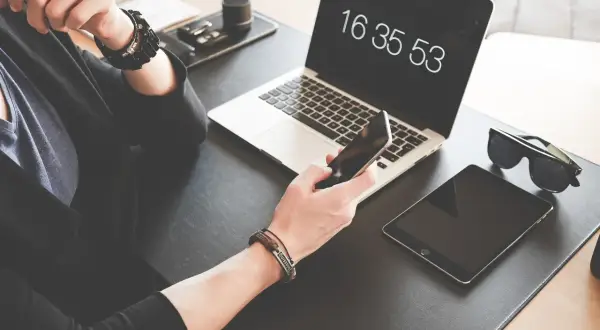 Osoba trzyma iPhone'a, obok leży tablet i laptop z widocznym zegarem.