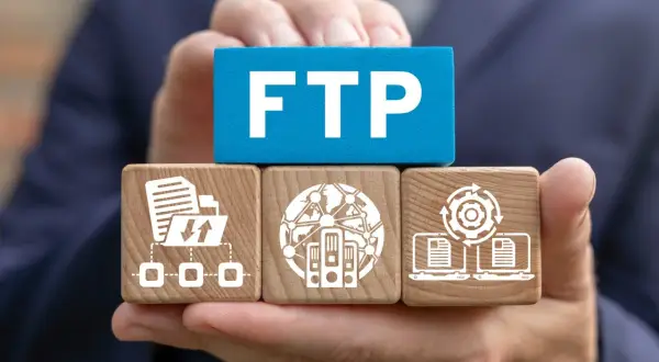 Ręce trzymają klocki symbolizujące transfer plików przez ftp: folder z ikonami pobierania/wysyłania, serwery w sieci, laptopy z dokumentami i mechanizmem.