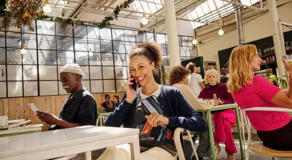 Uśmiechnięta kobieta rozmawia przez telefon, korzystając z eSIM T-Mobile. W tle ludzie w kawiarni.