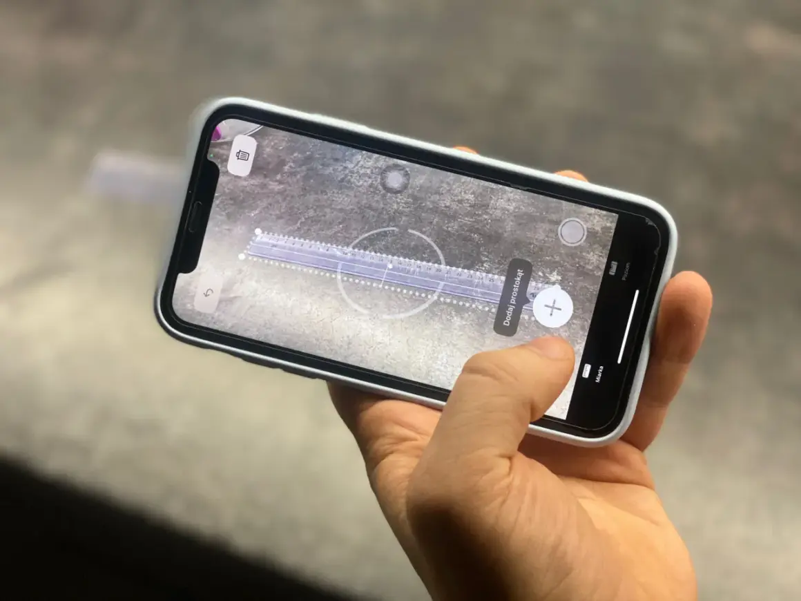 Dłoń trzyma smartfon, na ekranie którego działa aplikacja miarka. Wirtualna linijka mierzy odległość na powierzchni.