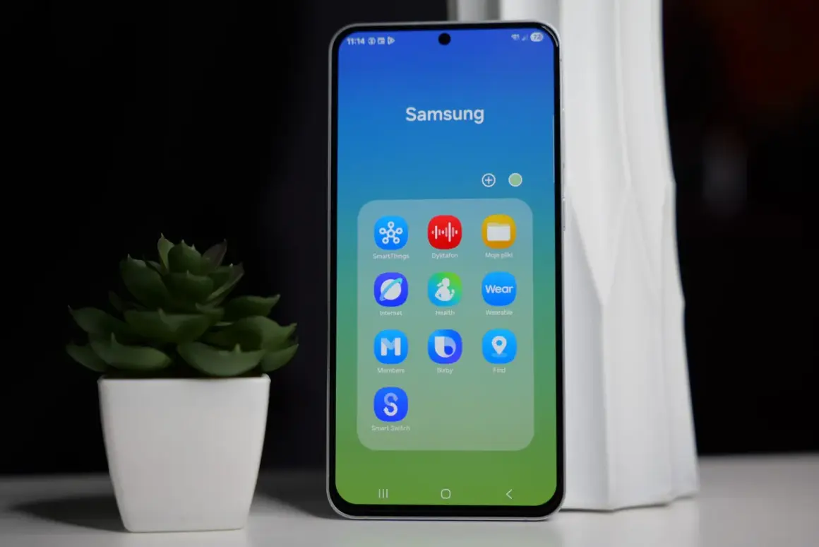 Telefon Samsung z ikonami aplikacji, w tym Bixby. Obok stoi zielona roślina w białej doniczce.