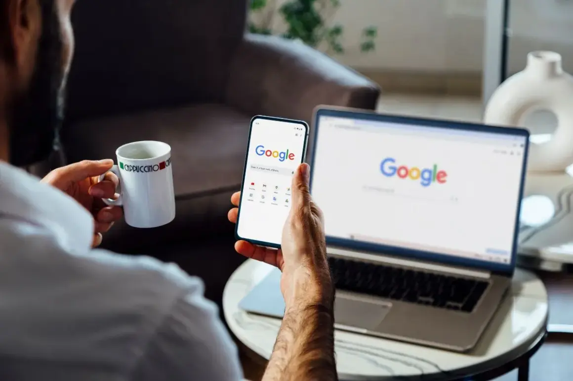 Mężczyzna z kubkiem kawy przegląda telefon, na którym widać stronę Google. Obok stoi laptop z otwartą wyszukiwarką. Chce wiedzieć, jak wylogować się z konta Google.