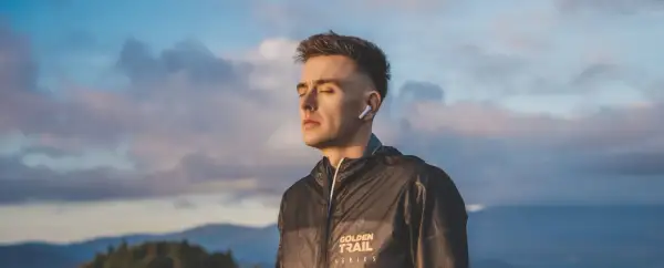 Młody mężczyzna w słuchawkach ANC, z zamkniętymi oczami, w kurtce z napisem "GOLDEN TRAIL SERIES", na tle nieba i gór.