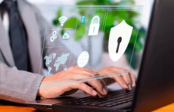 Osoba pracuje przy laptopie, a na ekranie wyświetla się interfejs z ikonami symbolizującymi cyberbezpieczeństwo: globus, mapa świata, chmura, kłódka i tarcza.