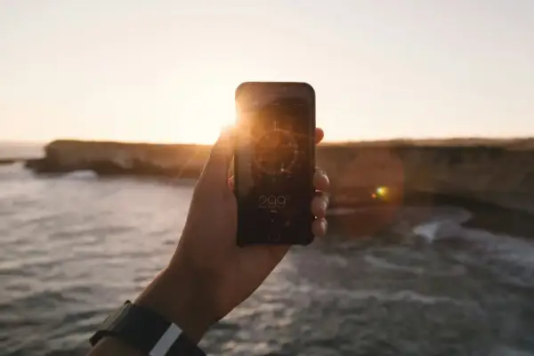 Dłoń trzyma telefon z włączonym kompasem w tle zachodzącego słońca nad morzem.